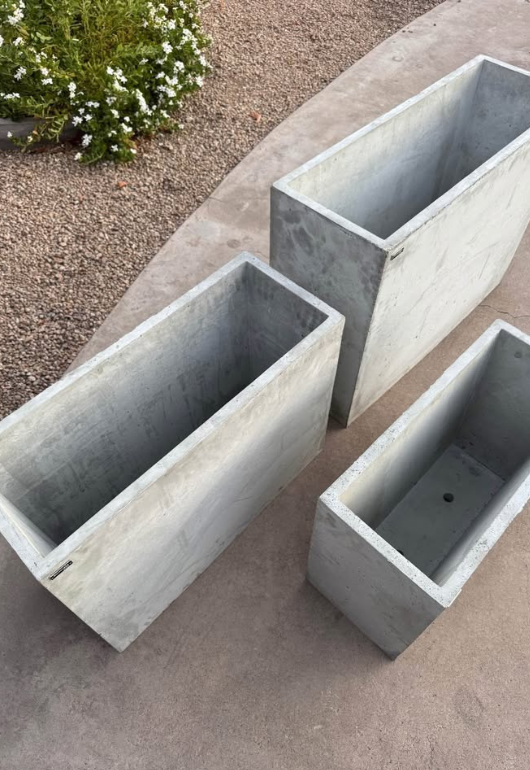 Three rectangular concrete planters on a concrete surface with a small bush in the background.
